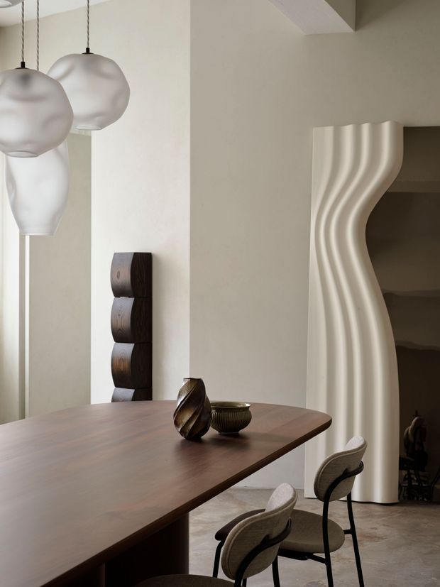 A refined dining area with a large dark timber dining table and upholstered dining chairs. Sculptural pendant lights hang above the table, while a dramatic undulating cream-colored architectural partition frames an opening to a darker adjacent space. Neutral plaster walls, warm wood, sculptural decor, and soft natural light create an elegant contemporary atmosphere.