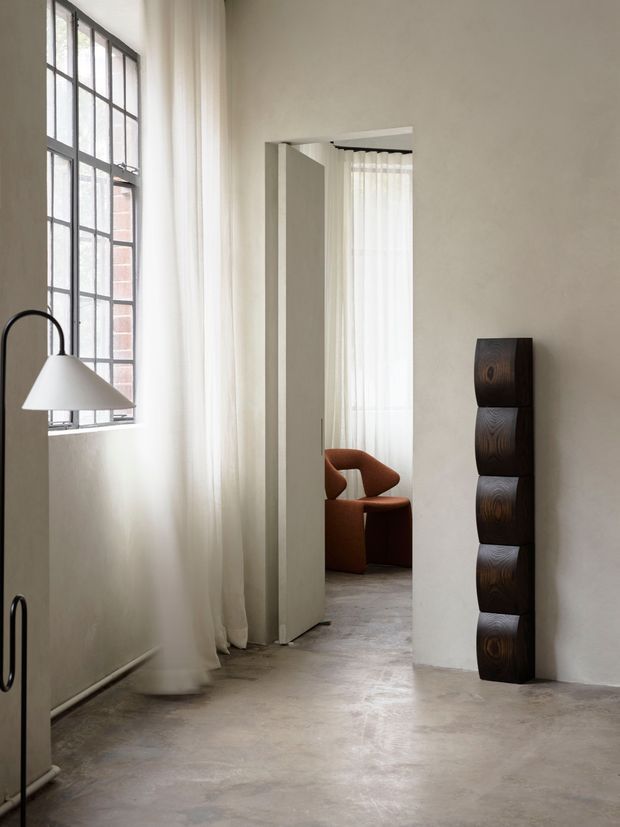 A minimalist interior passage or living area with tall industrial-style black-framed windows, sheer full-height curtains, pale plaster walls, and a polished concrete floor. A sculptural black floor lamp and stacked dark wooden decorative column add contrast. Through the open doorway, a rust-coloured sculptural lounge chair is visible in an adjoining room.