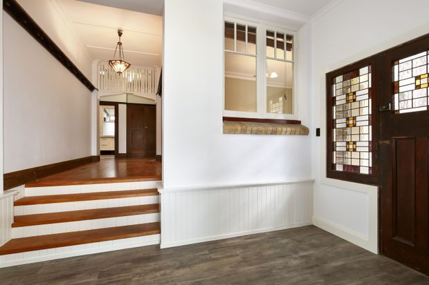 Traditional residential entryway with a dark timber stained-glass front door, matching stained-glass sidelight, white walls and trim, timber stair treads, and a wood-look floor. A raised hallway is visible beyond the short staircase, with decorative ceiling detailing, pendant lighting, and a timber-framed internal window with a projecting sill.