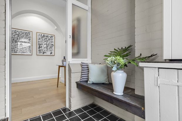 Bright entryway of a renovated brick building, featuring a black tiled floor, white-painted brick walls, a built-in dark timber bench with cushions and a vase of greenery, and a glazed internal door opening to a light timber-floored hallway with an arched opening and framed artwork.