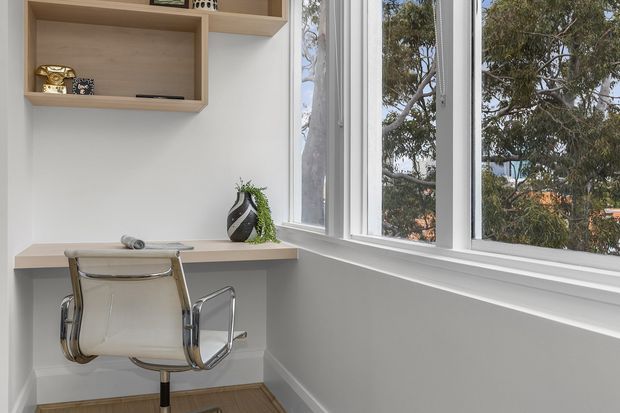 A compact, bright home office nook with a floating light-wood desk, a modern chrome-framed chair, built-in open shelving, and large white-framed windows overlooking leafy trees. The space has a clean, minimal, neutral aesthetic with strong natural light.