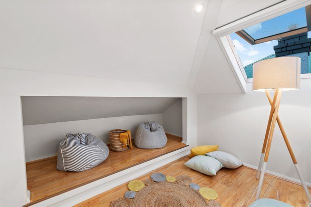 Bright attic-style sitting area with a built-in timber daybed or display platform beneath a sloped ceiling. The white room has a large skylight or roof window, warm timber flooring and platform, soft striped beanbags, cushions, a round woven rug, and a tripod floor lamp. A small portion of the exterior roofline is visible through the skylight.