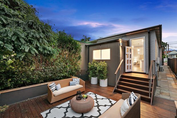 A compact contemporary dwelling with dark vertical cladding, a low-pitched roof and a raised entry accessed by timber steps. The image focuses on a warmly furnished timber deck courtyard, with outdoor seating, patterned rug, potted plants and dense screening vegetation. The open glazed door reveals a bright interior, while a paved side walkway runs alongside the building.