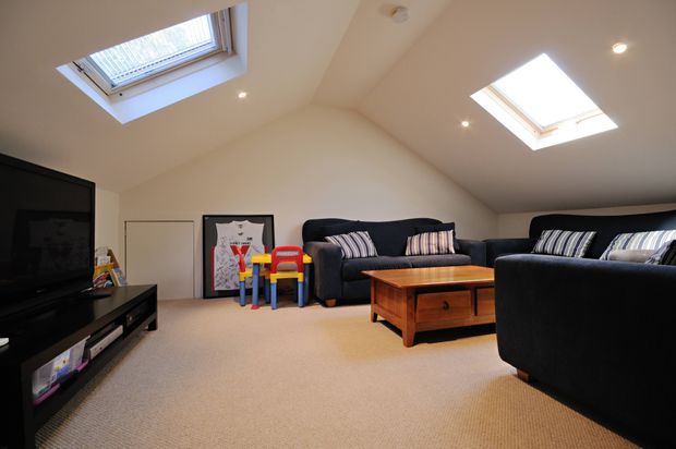 A bright attic-style family lounge or media room with a sloped ceiling, two skylights, recessed lights, beige carpet, dark sectional sofas, a wooden coffee table, and a television on a low media console. Children's chairs and toys are visible near the back wall.