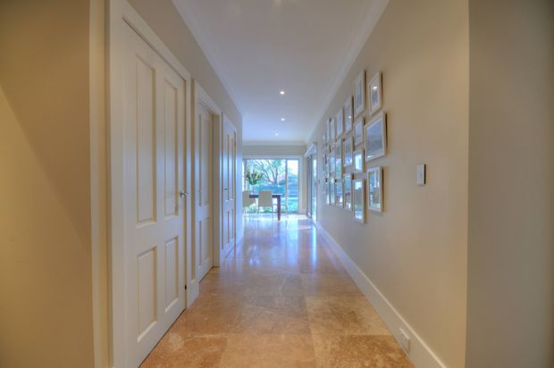 A long, bright residential hallway with warm beige walls, white raised-panel doors and trim, a polished natural-stone or marble-look floor, recessed ceiling lights, and a gallery wall of framed artwork. The corridor leads toward a glazed opening with a glimpse of a dining area and greenery beyond.