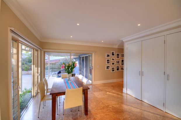 Warm, light-filled dining area with a rectangular timber dining table and pale upholstered chairs beside extensive glazed doors opening to an outdoor patio. The room has honey-toned walls, polished warm-toned flooring, white built-in floor-to-ceiling storage with shaker-style doors, recessed ceiling lights, decorative cornices, and a gallery wall of framed photographs. The space connects visually to a landscaped outdoor area through the glazing.