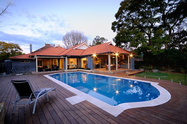 A large suburban house viewed from an expansive timber pool deck at dusk. The home has a warm terracotta tiled roof with multiple pitched roof sections, broad glazed openings, and a covered outdoor entertaining area supported by posts. A curved swimming pool occupies the foreground, surrounded by pale paving and dark timber decking, with lounge chairs and established trees around the property.