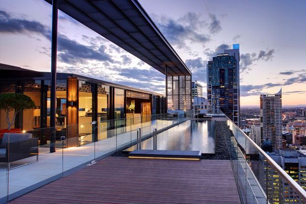 A sophisticated rooftop terrace overlooking a dense city skyline at dusk. The space features a long narrow reflecting pool or infinity-edge pool, timber decking, clear glass balustrades, a sleek covered outdoor lounge or dining area, warm architectural lighting, and contemporary metal framing.