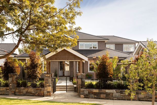 Exterior view of a large two-storey suburban house with a multi-gabled dark tiled roof, brown shingle-style wall cladding, light trim, and a covered entry porch. Stone retaining walls, black metal fencing, paved paths, and established planting frame the front facade.