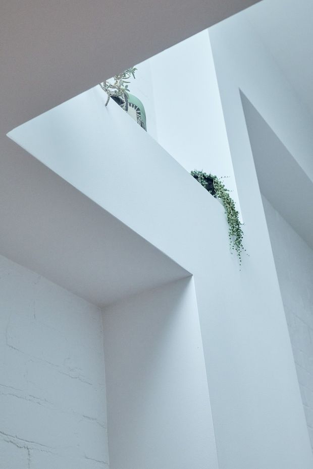 A minimalist white architectural interior viewed upward, featuring strong angular planes, a void or stair-like opening, and high-level ledges with trailing indoor plants. The space is bright and sculptural, with smooth white surfaces and subtle textured masonry visible on the lower wall.
