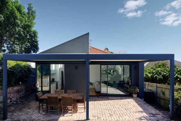 A contemporary single-storey residential extension viewed from an outdoor paved courtyard. A dark steel-framed covered terrace or carport-like structure spans the facade, with large glazed sliding doors opening into the interior. The courtyard is paved in warm brick, with timber outdoor dining furniture, garden planting, boundary fencing, and portions of a pitched terracotta-tiled roof visible behind the modern addition.