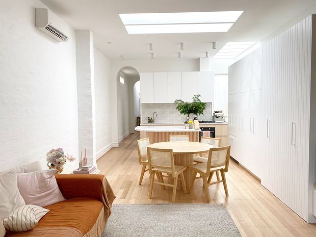 Bright open-plan kitchen and dining area with a small living zone. The room features white walls and floor-to-ceiling integrated cabinetry, pale timber flooring, a compact island or peninsula with timber detailing, a round timber dining table with matching woven-back chairs, and a rust-orange sofa. Large rectangular skylights and white linear ceiling lights create an airy, contemporary interior.