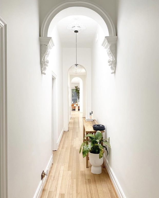 Bright, narrow residential hallway with white walls and a pale timber floor. Repeated arched openings, ornate plaster corbels and ceiling detailing create a refined traditional architectural character. A long timber console with a potted plant and small decorative objects runs along one wall, with pendant lights aligned down the corridor and a glimpse of a furnished room at the far end.
