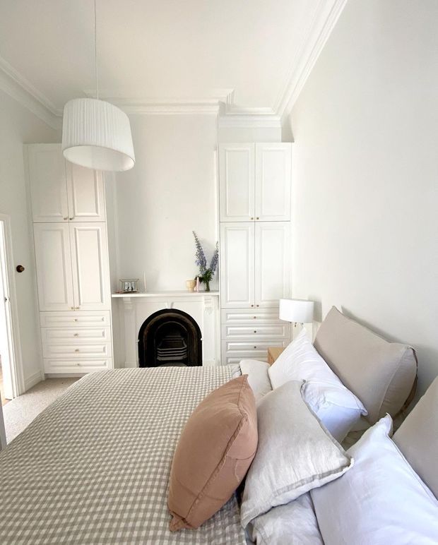 Bright, elegant bedroom with a large upholstered bed in the foreground, layered neutral bedding, extensive white built-in cabinetry, a decorative black fireplace with white mantel, classic ceiling cornices, and a white pendant light. The room has a soft, calm palette of white, cream, beige, and blush tones with traditional detailing.