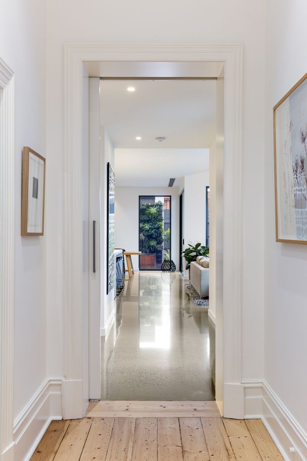 Bright view through a wide, heavily moulded interior doorway into a polished open-plan living area. The foreground has pale painted timber floorboards, while the adjoining space has a glossy speckled hard floor, white walls, recessed ceiling lighting, framed artwork, indoor plants, and glimpses of contemporary seating and dining furniture. A glass door or window at the far end opens toward greenery and neighbouring buildings.