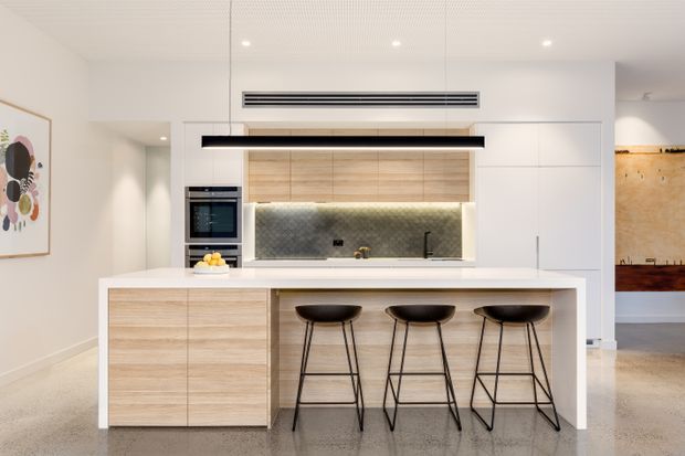 A bright contemporary kitchen centred on a large island with three black bar stools. The kitchen features white full-height cabinetry, light timber-grain cabinetry, a pale stone island benchtop, integrated appliances, a textured grey splashback, concealed rangehood, and a long black pendant light. A partial adjacent living or dining area is visible to the right.