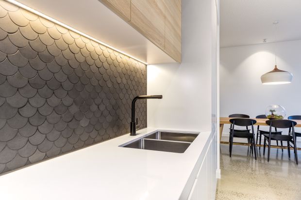 A contemporary kitchen focused on a white benchtop, stainless-steel undermount sink, matte black gooseneck tap, and a distinctive dark scalloped tile splashback. Light timber overhead cabinetry and integrated under-cabinet lighting frame the kitchen. Through the opening is a compact dining area with a timber table, black chairs, pendant light, and a polished concrete or terrazzo-like floor.