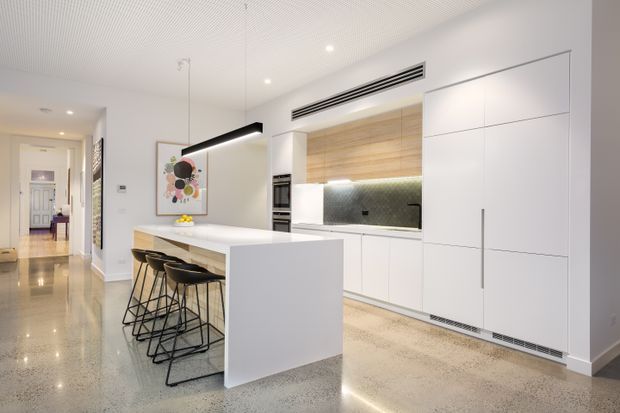 Bright contemporary open-plan kitchen with a large white waterfall-edge island, integrated white cabinetry, built-in appliances, timber-lined open shelving, and a dark textured splashback. Black pendant lighting and bar stools provide contrast, while polished light terrazzo or concrete-look flooring and a white linear ceiling create a clean, refined interior.