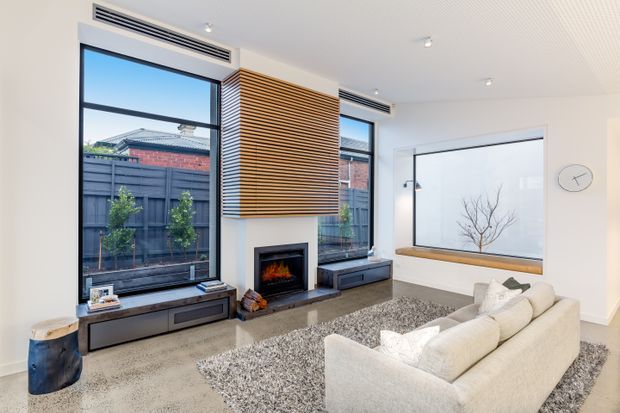 Bright contemporary living room with large black-framed windows, a fireplace with horizontal timber slat feature cladding, built-in low dark cabinetry, a broad frosted-glass feature window with timber sill, and a pale sectional sofa over a textured grey rug. The room has a clean, refined fit-out with integrated linear air vents and recessed lighting.