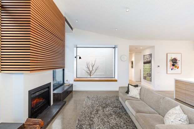 Bright contemporary living room with a large L-shaped light-grey sectional sofa, textured area rug, built-in fireplace, and a timber slatted feature wall extending across the ceiling-side volume. White walls, recessed lighting, a large window with a built-in timber sill, minimalist artwork, and timber-toned cabinetry create a warm, refined interior.