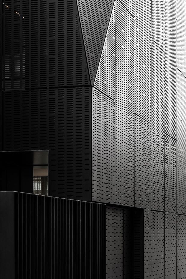 A dramatic close-up architectural exterior showing tall, angular building surfaces clad in dark perforated metal panels. The facade features dense rectangular perforations, strong vertical and diagonal geometry, deep shadows, and a monochromatic black and charcoal palette. A dark metal balustrade and recessed opening appear at the lower left, emphasizing the building's contemporary urban character.