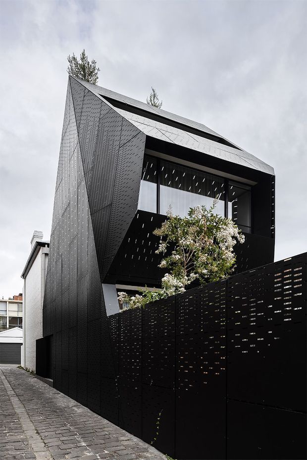 Street-level exterior view of a narrow contemporary multi-storey residence in a dense suburban setting. The house has a sharply folded, angular gabled roof and facade clad in dark perforated metal panels, with a recessed upper-level glazed opening and a perforated privacy screen enclosing the front boundary. A paved lane and neighbouring buildings are visible.