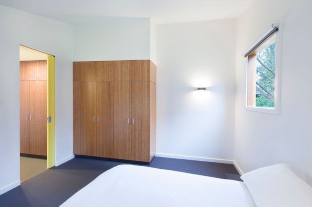 Bright, uncluttered bedroom with white walls and ceiling, dark carpet, and a built-in warm timber wardrobe. A partially open doorway reveals matching timber joinery in the adjacent space, while a window provides natural light and views of greenery. The lower foreground shows part of a white bed and pillows.