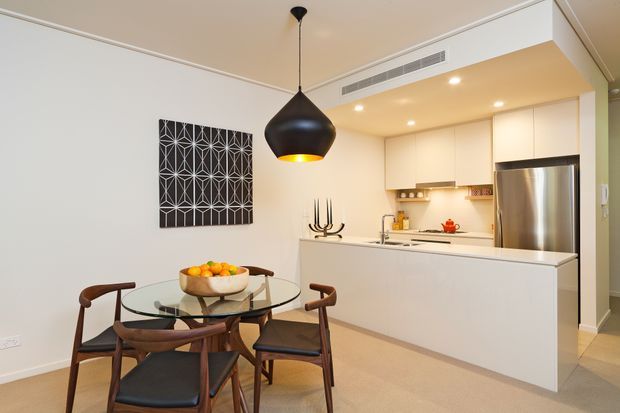 A bright open-plan dining and kitchen area with a round glass dining table and four dark timber chairs in the foreground. The compact kitchen features white flat-panel cabinetry, a long white island, stainless-steel refrigerator, integrated cooktop and rangehood, and warm recessed lighting. A large black pendant light and geometric black-and-white wall artwork provide contrast against the neutral walls and light flooring.