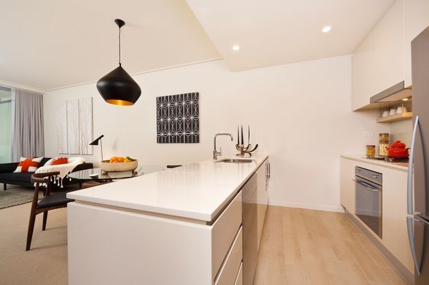 Bright open-plan kitchen with a large white island and integrated sink, pale timber flooring, white walls and cabinetry, stainless-steel appliances, and a black pendant light. A compact living area with sofa seating is visible beyond the kitchen.