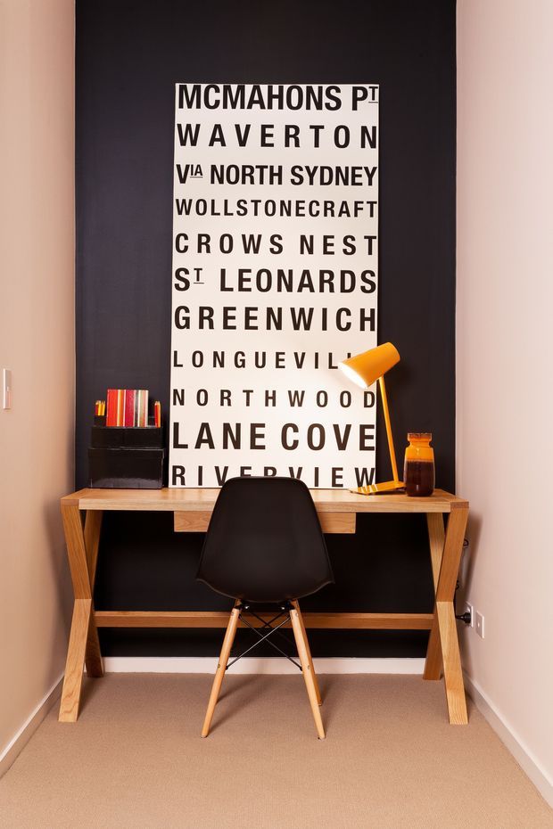 A compact home office nook with a timber desk positioned against a black feature wall. The desk is paired with a black molded chair, an orange task lamp, a small black storage unit with colourful books, and a large typographic city-name artwork. Warm neutral walls and carpet create a cosy, contemporary workspace.