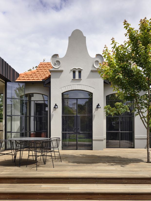 Exterior view of a characterful rendered building opening onto a timber deck and dining terrace. The facade features a highly decorative curved gable with circular ornamental details, arched brick detailing above tall black-framed steel-look windows and doors, and a terracotta tiled roof section. A contemporary outdoor dining setting sits in the foreground, with mature planting beside the deck.