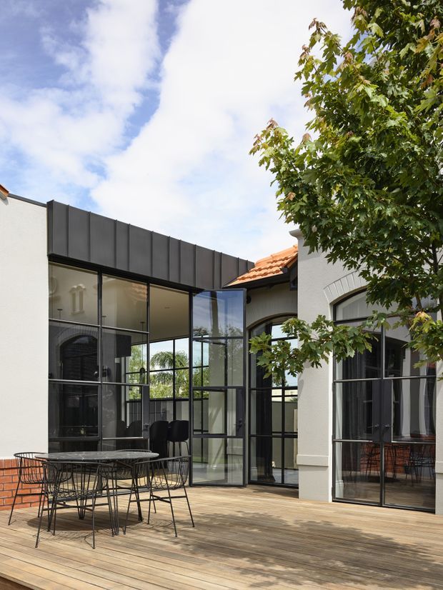 A bright courtyard-style exterior showing a timber deck with a black wire-frame outdoor dining table and chairs, surrounded by white rendered walls and extensive black-framed floor-to-ceiling glazing. The addition has a dark metal-clad roof edge and connects to an older-looking rendered building with arched openings and terracotta roof tiles. A leafy tree partially frames the scene.