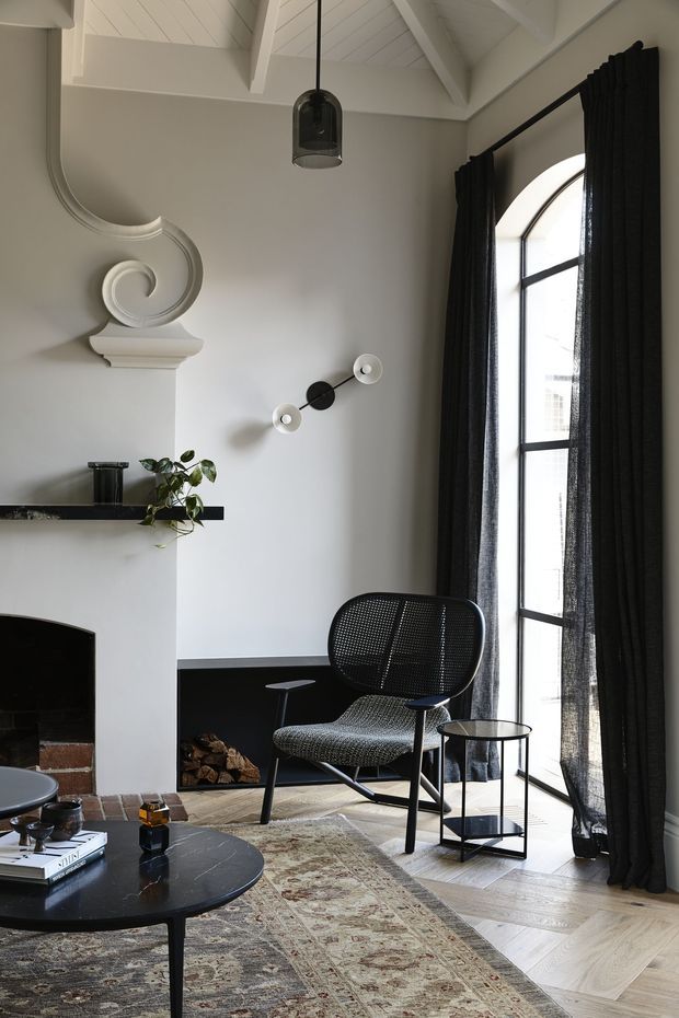 A refined living room with a tall arched black-framed window, full-height black curtains, white-painted vaulted timber ceiling with exposed beams, and a sculptural decorative fireplace mantel. The room features a black marble coffee table, woven lounge chair, small side table, patterned rug, dark fireplace surround, and restrained contemporary furnishings.