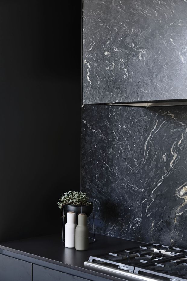 Close-up of a dark, sophisticated kitchen featuring a black benchtop and cabinetry, a dramatic dark veined stone splashback, and a stainless-steel gas cooktop. Minimal decorative vases sit beside the cooktop, with black and charcoal tones dominating the composition.