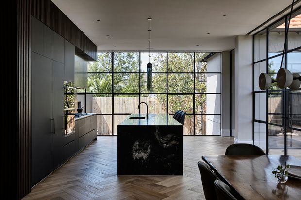 A refined open-plan kitchen and dining area with dramatic floor-to-ceiling black-framed glazing overlooking trees and a timber fence. The kitchen features dark full-height cabinetry, a black stone island with an integrated sink and sculptural pendant lighting, while the dining table occupies the foreground. Herringbone timber flooring and restrained contemporary detailing create a sophisticated, moody interior.