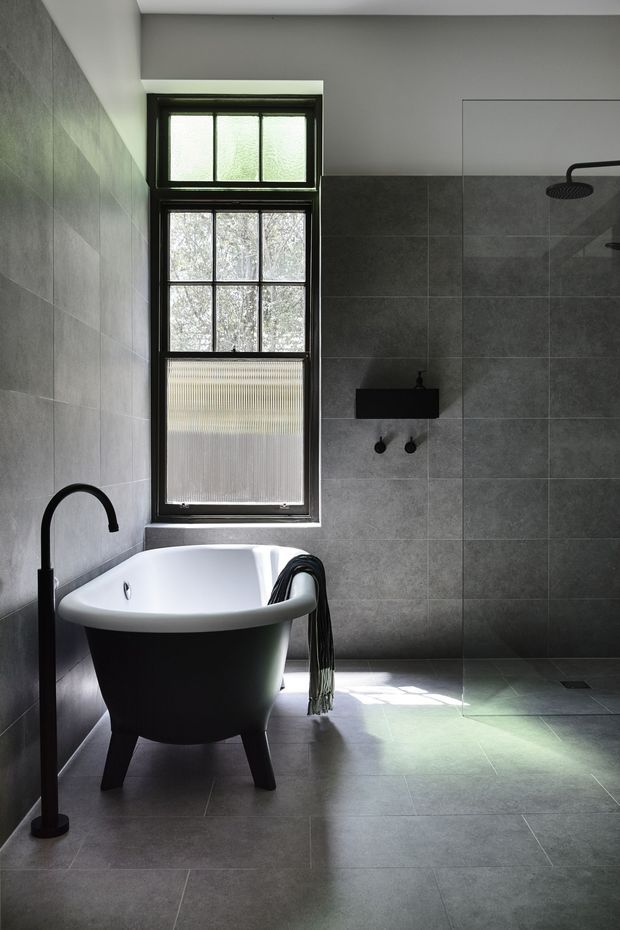 A dark, refined bathroom featuring a freestanding white bathtub with a black exterior, matte black floor-mounted tap, wall-mounted shower fittings, and a frameless glass shower screen. Large-format grey stone-look tiles cover the walls and floor, complemented by black-framed glazing and a high narrow window bringing in filtered daylight.