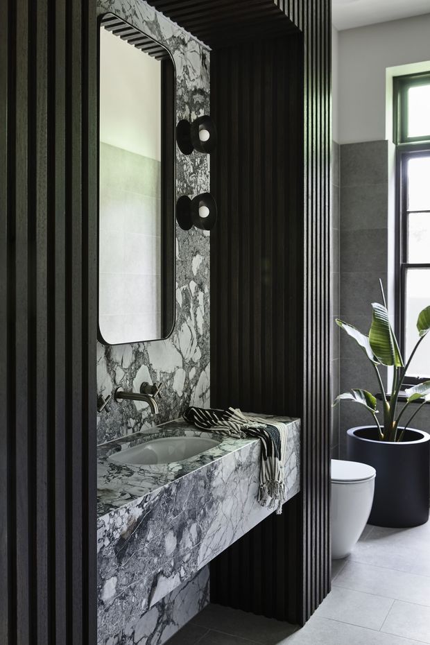 A sophisticated contemporary bathroom featuring a floating vanity clad in dramatic veined grey marble, a matching marble splashback and integrated basin, a rounded rectangular mirror, black wall-mounted tapware, and black vertical fluted wall panelling. A toilet, large potted plant, grey tiled floor, and dark-framed window are visible to the right. The palette is dark, monochromatic, and luxurious with natural light from the window.
