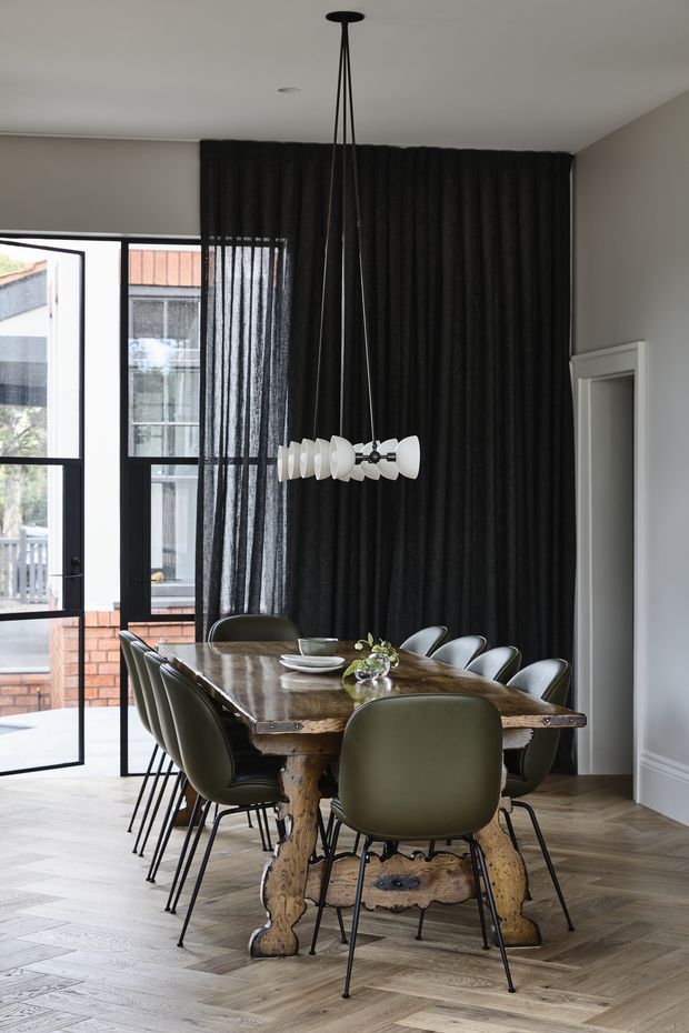 A refined dining room with a large rustic timber dining table surrounded by olive-green upholstered chairs. The room features herringbone timber flooring, a dramatic black full-height curtain, black-framed glazed doors and windows, white walls and skirting, and a sculptural modern pendant light suspended above the table. An adjacent exterior courtyard or balcony is visible through the glazing.