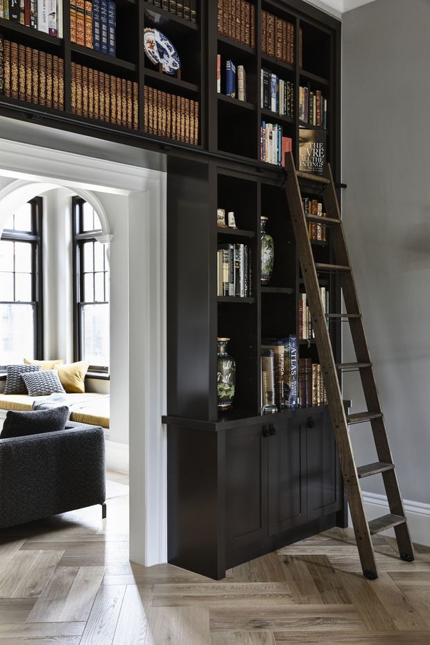 A refined home library with full-height dark built-in bookcases filled with books and decorative objects, lower enclosed cabinetry, and a leaning wooden library ladder. The room has dark grey walls, white architectural trim, herringbone timber flooring, and a wide arched opening leading to a bright sitting area with tall windows and upholstered seating.