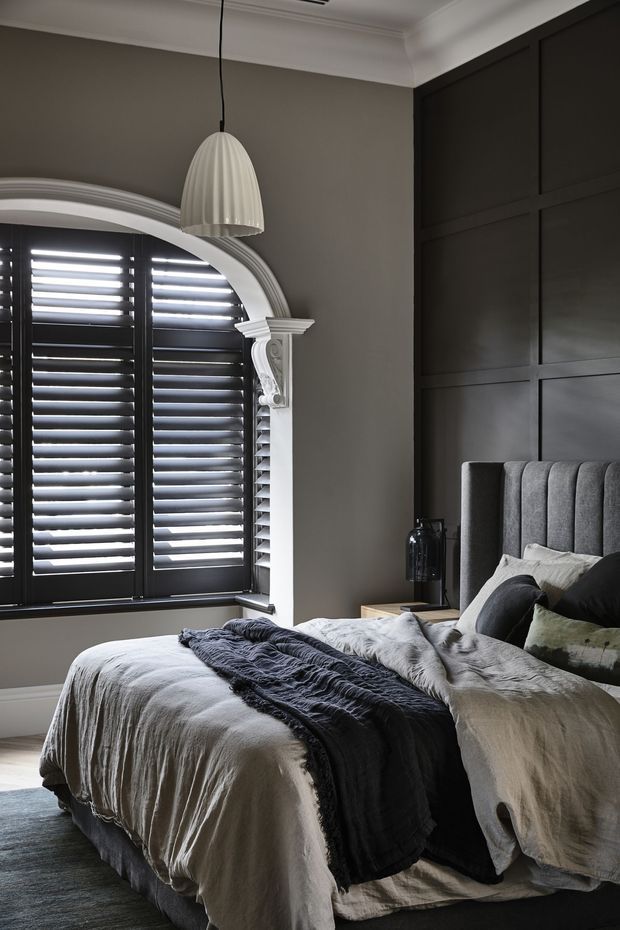 A sophisticated bedroom with a large upholstered bed dressed in layered grey and off-white linens, a dark charcoal throw, and assorted cushions. The room features a dramatic dark timber wall with geometric paneling, a grey-painted wall, a decorative arched window with white moulded trim and black plantation shutters, and a sculptural pendant light. The palette is moody, refined, and monochromatic with traditional architectural detailing.