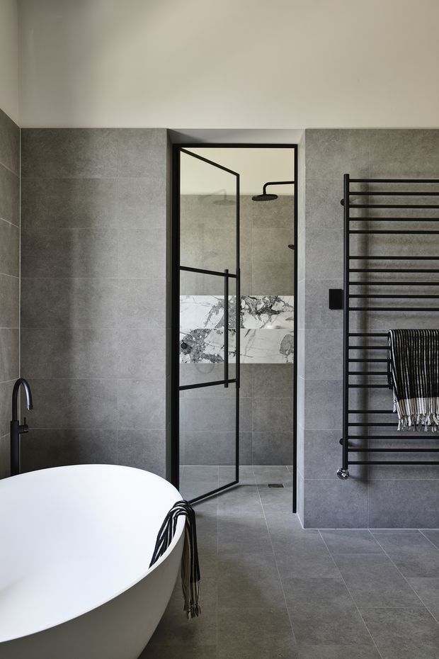A refined contemporary bathroom with large grey tiled walls and floor, a freestanding white bathtub in the foreground, and a black-framed glass shower enclosure. A marble-look feature band runs through the shower, complemented by matte black tapware, shower fittings, and a black heated towel rail.