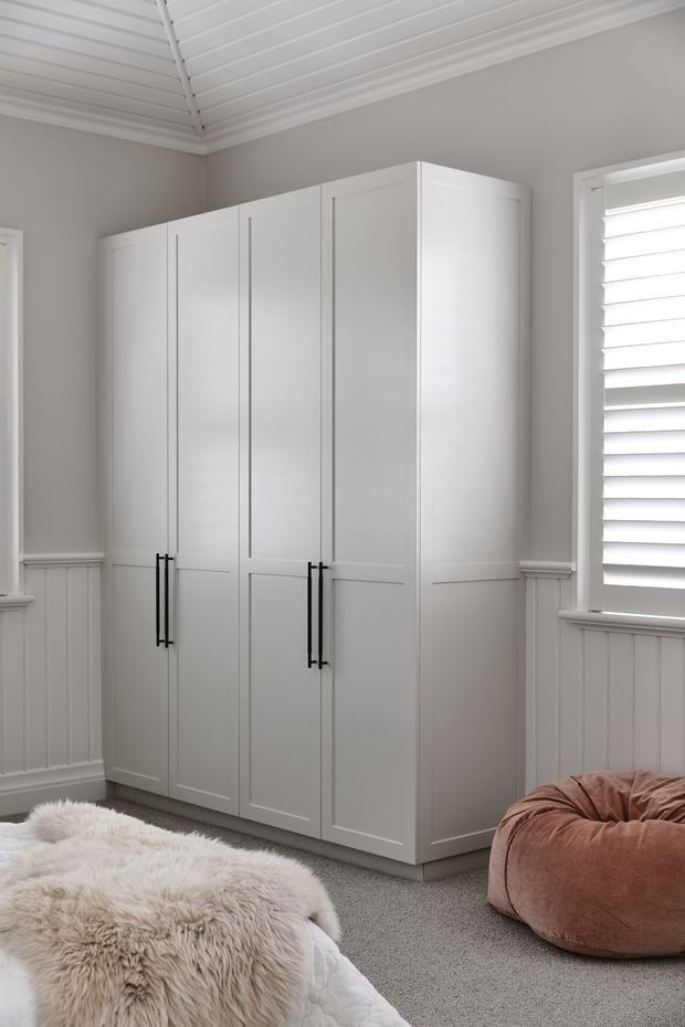 A bright, softly styled bedroom corner featuring a large built-in white wardrobe with paneled doors and black vertical handles. The room has pale grey walls, white decorative wall panelling, a white slatted ceiling, plantation shutters, and grey carpet. A bed with a plush cream sheepskin throw and a muted blush beanbag are partially visible.