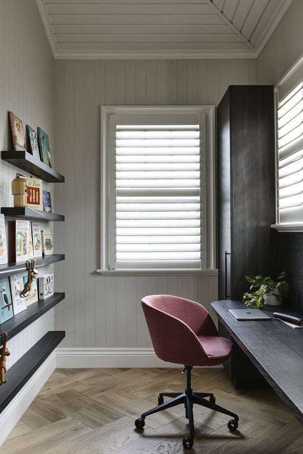 A compact, thoughtfully designed home office with vertical grey wall panelling, white plantation shutters, a herringbone timber floor, dark built-in shelving and cabinetry, and a dark desk beneath the window. A burgundy upholstered swivel chair, laptop, houseplant, and books add colour and function. The room is bright with a refined, tailored aesthetic.