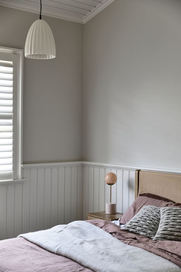 A softly styled bedroom corner with a made bed, upholstered beige headboard, mauve and grey bedding, and a small bedside table with a spherical lamp. The room features grey painted walls, white vertical wall panelling and trim, a white beadboard-style ceiling, and a window with white plantation shutters. The atmosphere is calm, muted, and refined.