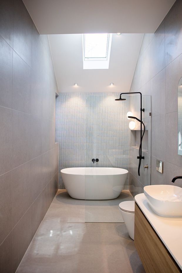 A narrow, contemporary bathroom with a freestanding white bathtub beneath a skylight, a walk-in shower with black fixtures and glass screen, and a timber-vanity cabinet with a white vessel basin. Large grey wall and floor tiles create a calm, streamlined finish, while a vertically ribbed feature-tile wall adds texture.