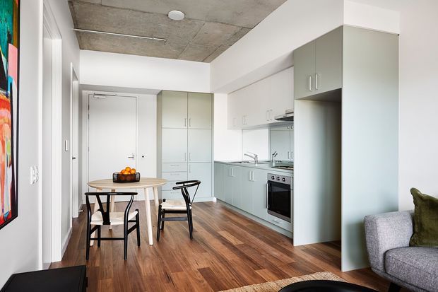 Compact open-plan apartment interior combining a small kitchen, dining area and living space. The room has white walls and cabinetry, pale grey-green joinery, warm timber flooring, a raw concrete ceiling, and black metal dining furniture. A partial sofa and colourful artwork are visible in the foreground.