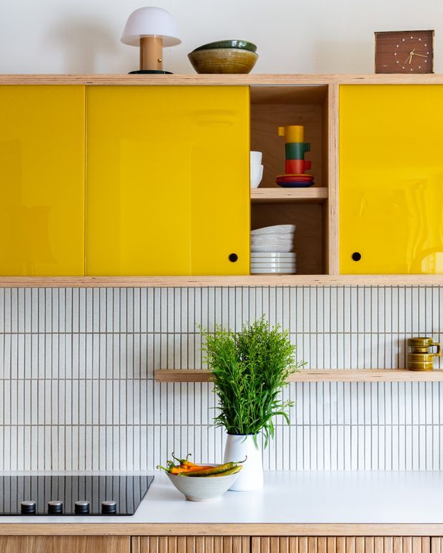 Close-up view of a cheerful retro-inspired kitchen with glossy mustard-yellow overhead cabinet doors, open timber shelving, and a vertically ribbed light tile splashback. The white benchtop includes a black induction cooktop, a bowl of fruit, and a potted plant, while timber shelving and a fluted timber base cabinet add warmth. Decorative mid-century objects sit atop the cabinetry.