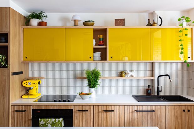 Bright contemporary kitchen with a warm timber-grain base cabinetry, glossy mustard-yellow upper cabinet fronts, vertically ribbed white tile splashback, white benchtops, black hardware and tapware, and open shelving with plants and everyday kitchenware. A cooktop, oven, sink and stand mixer are visible.