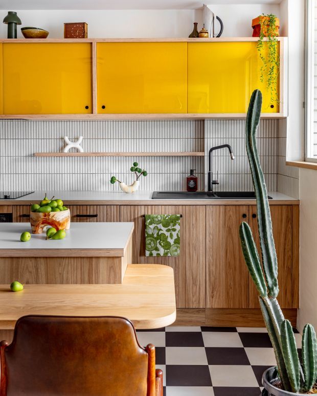 A stylish kitchen with warm timber cabinetry, bright glossy yellow overhead cupboard doors, a vertically lined tiled splashback, black sink and tapware, and a black-and-white checkerboard floor. A timber island with a light countertop and leather dining chair occupies the foreground, while decorative objects and tall indoor cacti add character.