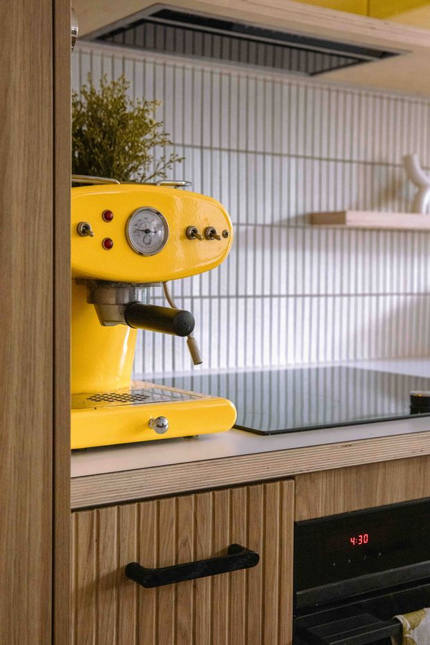 Close-up of a compact retro-inspired kitchen featuring a bright yellow espresso machine on a timber-look benchtop, a black induction cooktop, integrated oven, and vertically fluted timber cabinetry. The backsplash and upper wall are finished in narrow vertical white tiles, with open shelving and a small plant adding detail. A recessed ceiling vent or skylight is visible above.